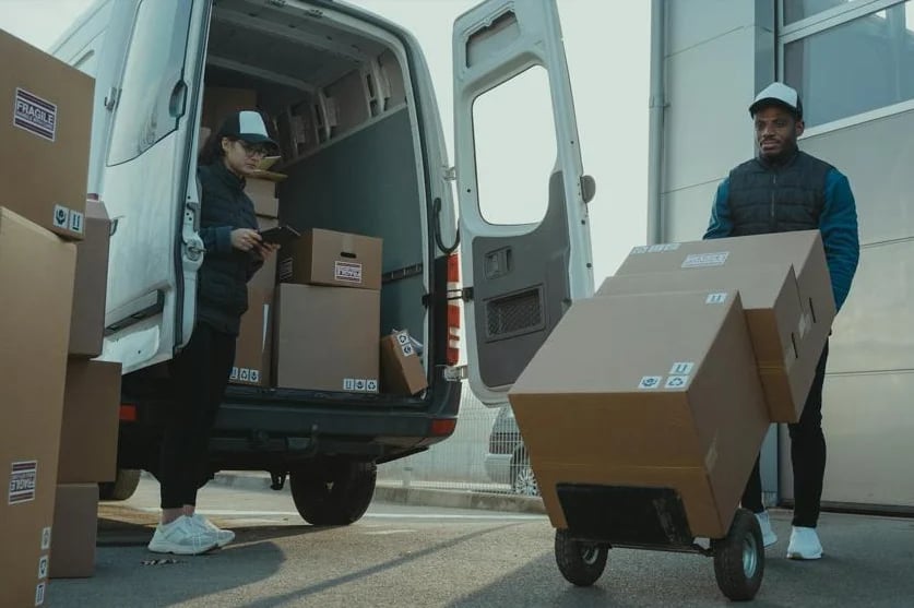 Two delivery workers unloading cardboard boxes from a white van onto a hand truck during a delivery operation